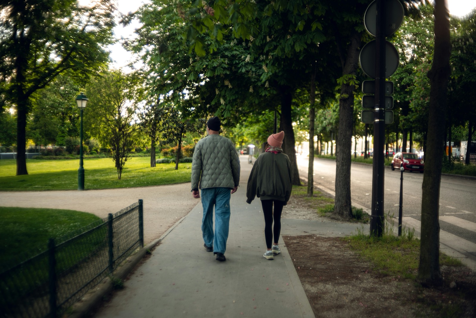 Two people walking side by side along a tree-lined sidewalk beside a park, viewed from behind.