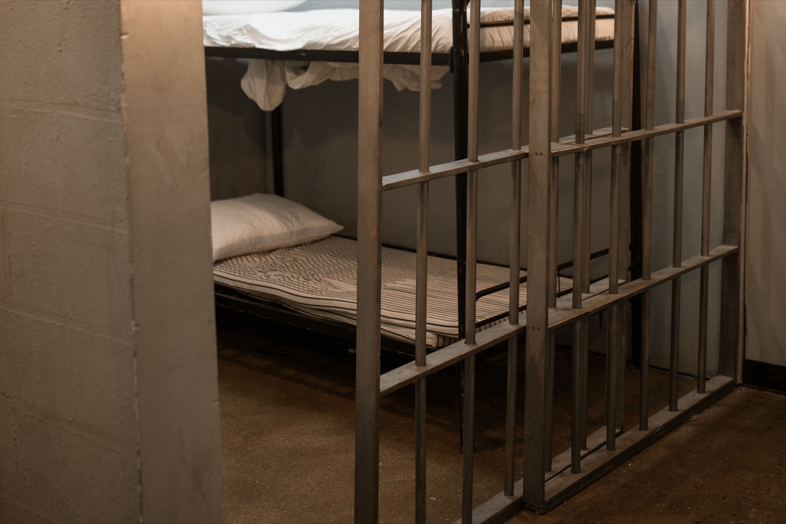 A jail cell photographed through steel bars, with a bare bunk bed, a thin striped mattress and a folded white blanket inside.